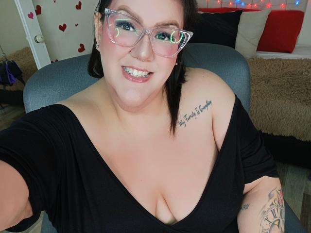 GiiannaMorrys with glasses, black off-shoulder top, showing her tits, tattoo on right shoulder, smiling, fair skin, black hair, arm tattoo, seated, looking at camera.