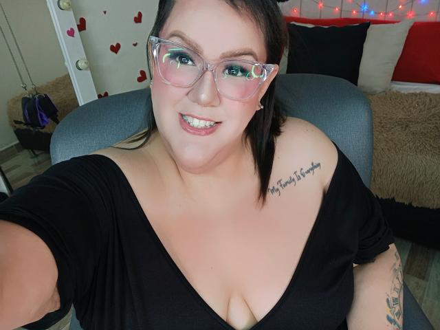 GiiannaMorrys with glasses, black top, showing cleavage, smiling, tattoo on right shoulder, black hair, tattoo on left arm, sitting, sexy, pale skin, small nose, green eyes.