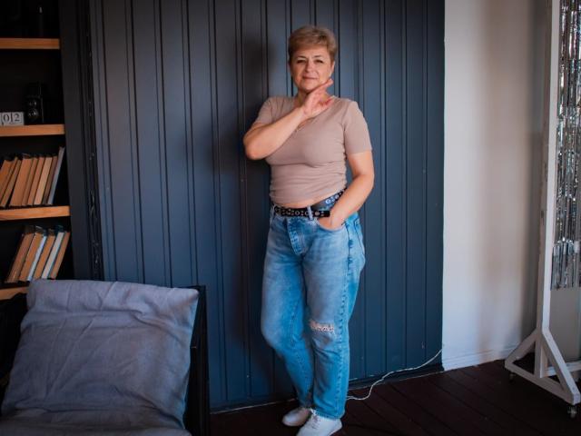 EmiliaGree with short blonde hair, tan t-shirt, and blue ripped jeans, standing against dark blue wall, hand near chin, wearing white socks, smiling slightly, in cozy room.