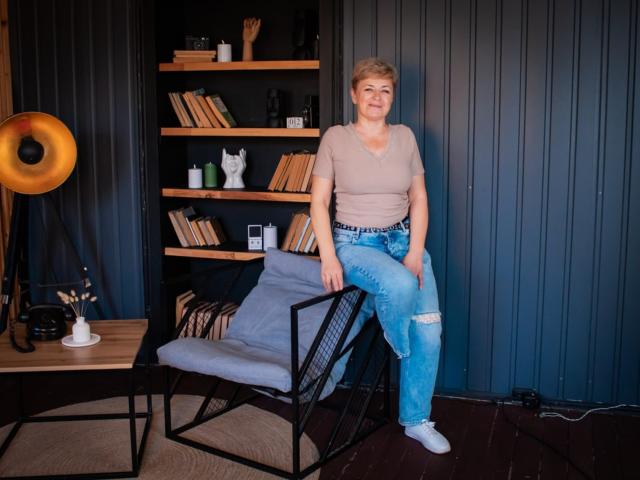 EmiliaGree with short blonde hair, gray V-neck shirt, ripped blue jeans, and white sneakers, standing on a black metal chair, in a room with a wooden shelf.