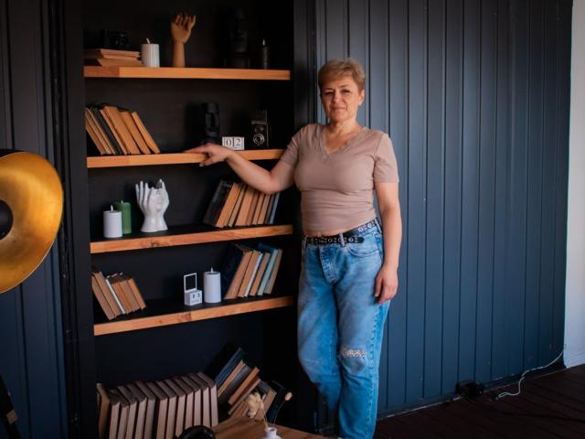 EmiliaGree stands in front of a shelf, wearing a beige top and ripped blue jeans, pointing at books with her right hand, showing a cute ass.