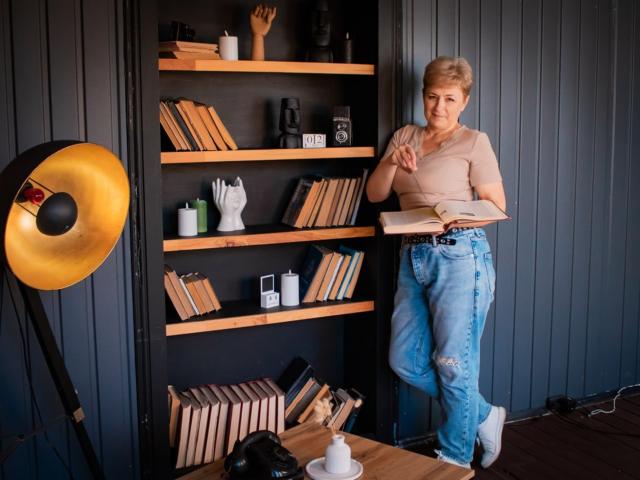 EmiliaGree stands in a room, wearing a beige top and ripped blue jeans, holding an open book, next to wooden shelves with books and a white hand-shaped ornament.