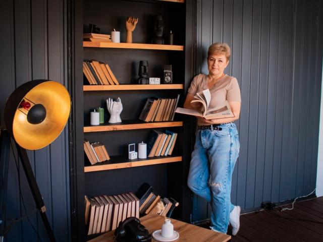 EmiliaGree with short blonde hair, beige top, ripped blue jeans, reading a book, standing in front of a black wall with wooden shelves, large gold studio light on left.