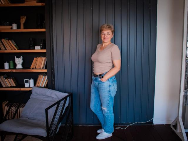 EmiliaGree stands with hands in blue jeans, wearing a beige top, white socks, and a belt, against a dark blue, ribbed wall, next to a gray chair.