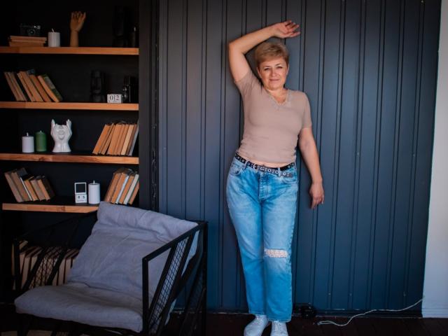 EmiliaGree, short blonde hair, beige top, ripped blue jeans, white socks, leaning against gray wall, arm raised, casual pose, bookshelf with white cat figurine on left.