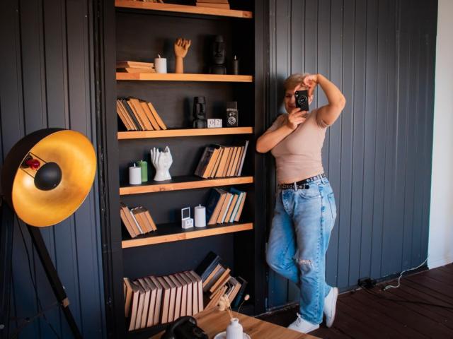 EmiliaGree with blonde hair, tan top, and blue jeans, taking a mirror selfie, standing by a black wall with wooden shelves, next to a gold studio light.