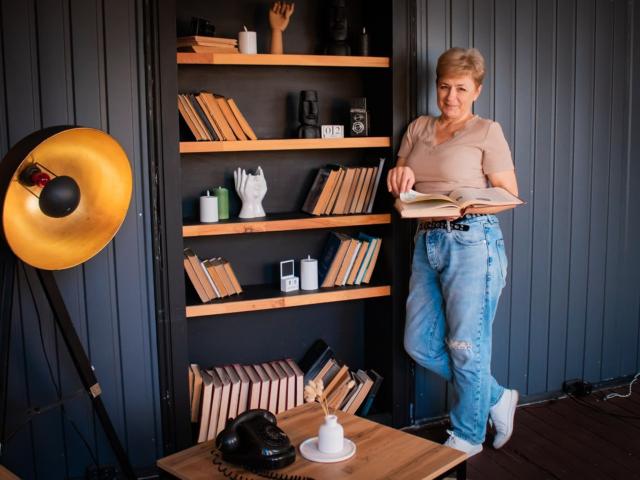 EmiliaGree stands reading, wearing a beige top, blue ripped jeans, white socks, holding a book. Shelves with books and decor behind her, a black lamp to the left.