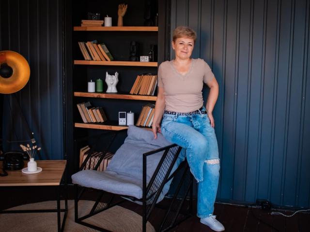 EmiliaGree with short blonde hair, tan shirt, ripped blue jeans, and white socks, sitting on a black metal chair, in a cozy room with wooden shelves and dark walls.