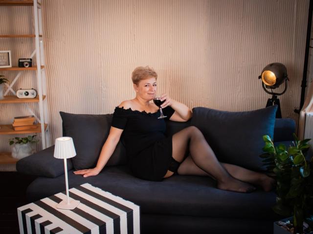 EmiliaGree, short blonde hair, black off-shoulder dress, black stockings, holding wine glass, sitting on gray sofa, leg up, lamp on striped table, plant, studio light in background.