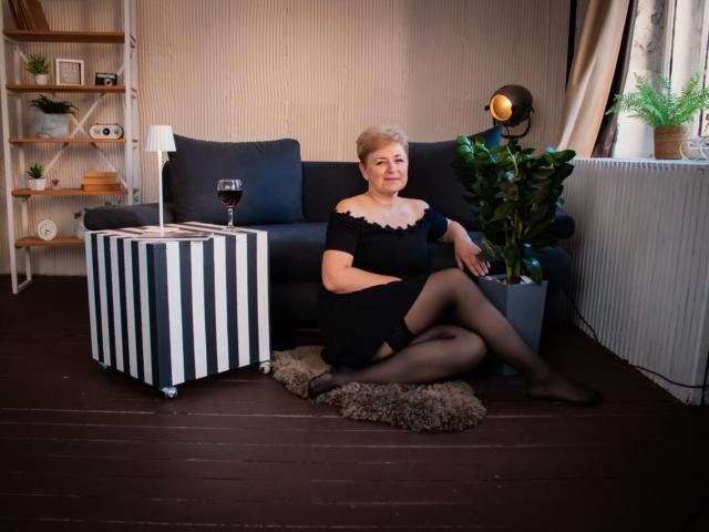 EmiliaGree sits on a fluffy rug, wearing a black dress, sheer stockings, and black heels, with a glass of red wine on a striped table. Her ass is visible.