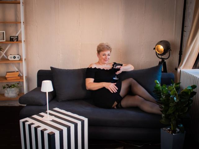EmiliaGree, blonde woman with short hair, wearing black off-shoulder dress and black stockings, sitting on dark gray sofa, holding a glass, legs crossed, lamp and plant nearby.