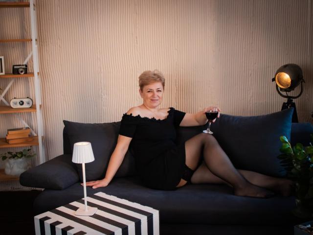 EmiliaGree with short blonde hair, black off-shoulder dress, black stockings, holding wine glass, sitting on dark gray couch, smiling, right arm on cushion, left leg crossed.