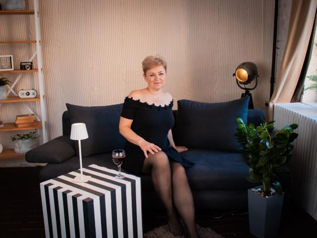 EmiliaGree with short blonde hair, black off-shoulder dress, black tights, sitting on dark blue couch, holding wine glass, striped black and white table, potted plant, lamp.