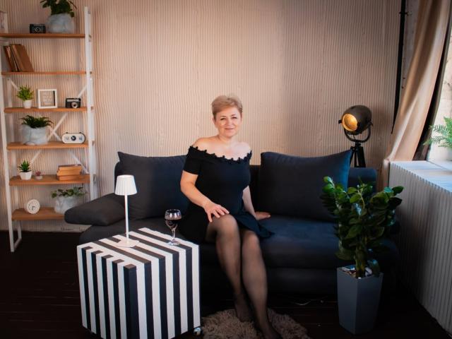 EmiliaGree sits on a dark blue couch, wearing a black off-shoulder dress, black stockings, and short blonde hair. Glass of wine on striped table, potted plant nearby.