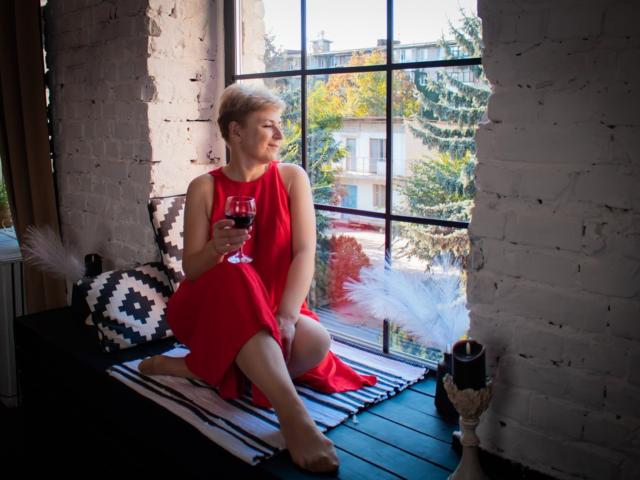 EmiliaGree with short blonde hair, wearing a red dress, sitting on a black and white striped rug, holding a glass of red wine, by a window, white brick wall.