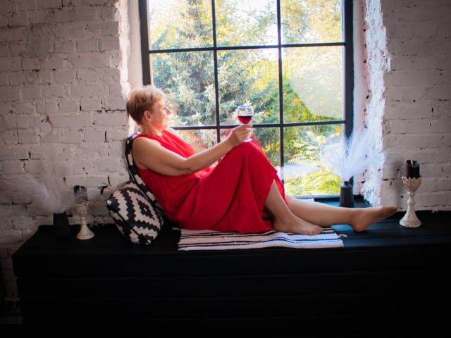EmiliaGree, short blonde hair, red dress, bare legs, sitting on black window ledge, holding glass of red wine, black and white pillow, white brick wall, sunlight.
