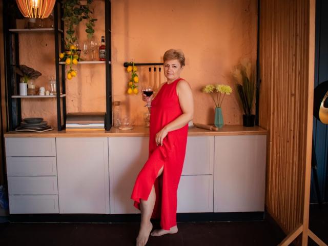 EmiliaGree in a red dress with a thigh slit, holding a glass of red wine, standing barefoot, blonde, cute, medium tits, in a warm, cozy room.