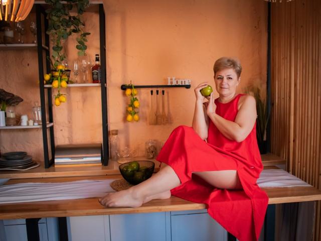 EmiliaGree with short blonde hair, wearing a red dress, sitting on a kitchen counter, holding a green apple, her bare feet on the wood, a bowl of apples nearby.