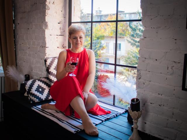 EmiliaGree with blonde hair, wearing a red dress, holding a glass of red wine, sitting on a black bench, white brick walls, black and white patterned pillow, and white feathers.