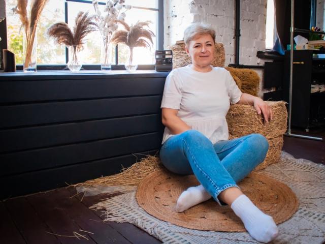 EmiliaGree with short gray hair, white shirt, blue jeans, white socks, sitting on hay, legs crossed, smiling, black dresser, windows, potted plants, wooden floor, rustic room.