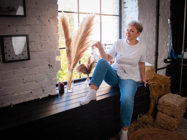 EmiliaGree with short blonde hair, white t-shirt, blue jeans, and white socks, sitting on black wooden ledge, touching pampas grass, with hay bales and white brick wall.