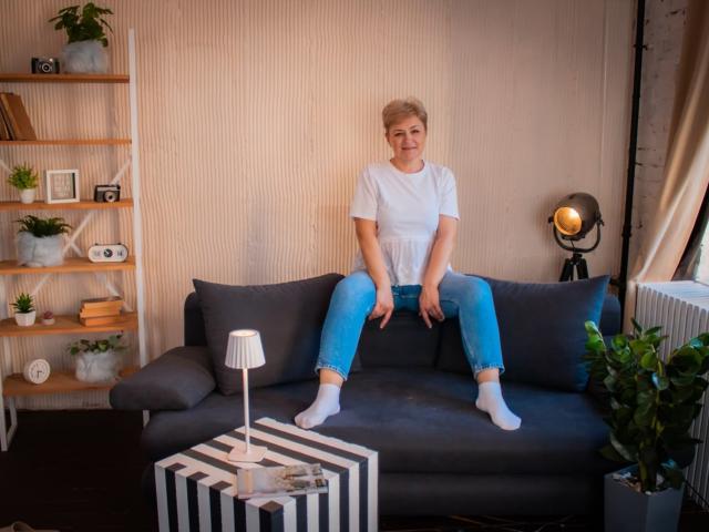 EmiliaGree with short blonde hair, white t-shirt, blue jeans, and white socks, sitting on dark gray couch, hands on thighs, smiling, in cozy living room.