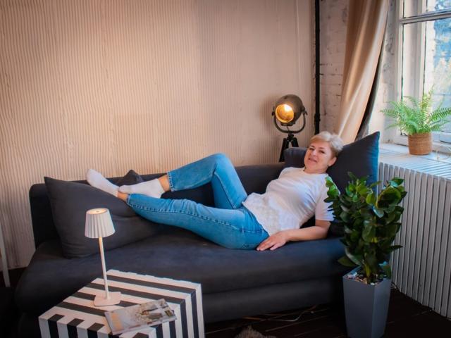 EmiliaGree with short blonde hair, white tee, blue jeans, white socks, lying on dark gray couch, legs up, smiling, small lamp, potted plant, window, and studio light in background.