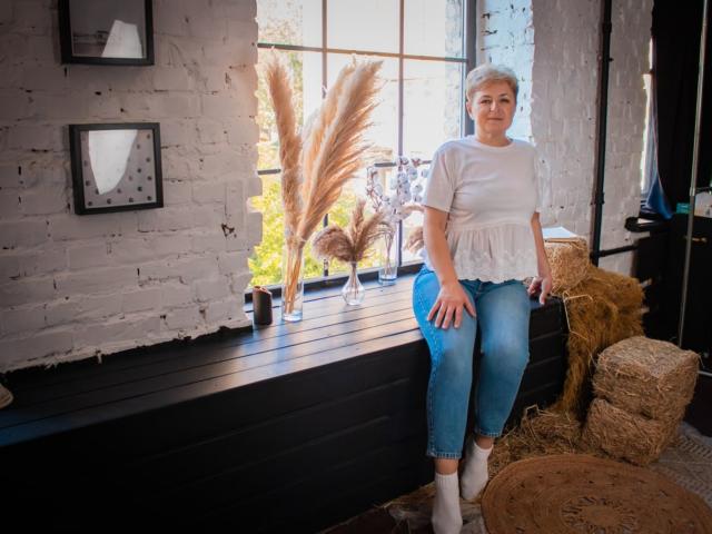 EmiliaGree with short gray hair, white shirt, blue jeans, and white socks, sitting on black wooden window ledge, surrounded by hay bales, pampas grass, and framed pictures.