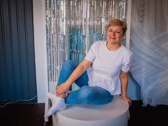 EmiliaGree with short blonde hair, white t-shirt, and blue jeans, sitting on a white stool, smiling, wearing white socks, with her legs crossed, in a casual setting.