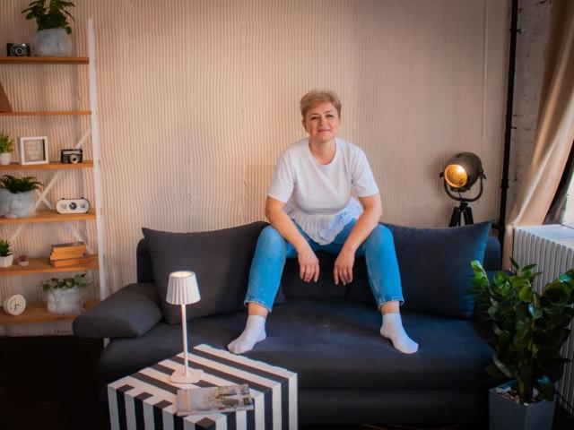 EmiliaGree, chubby with short blonde hair, wearing a white tee and blue jeans, sitting on a dark gray couch, legs spread, white socks, small smile, casual setting.