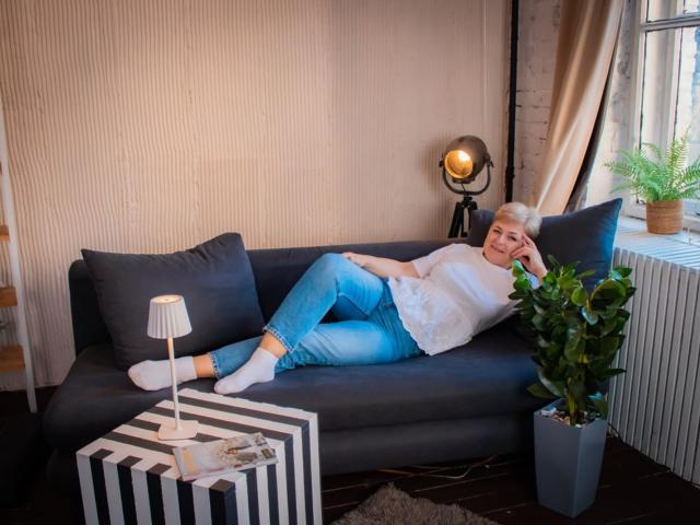 EmiliaGree with short gray hair, white turtleneck, blue jeans, and white socks, lounging on a dark blue sofa, legs crossed, next to a green plant and striped table.
