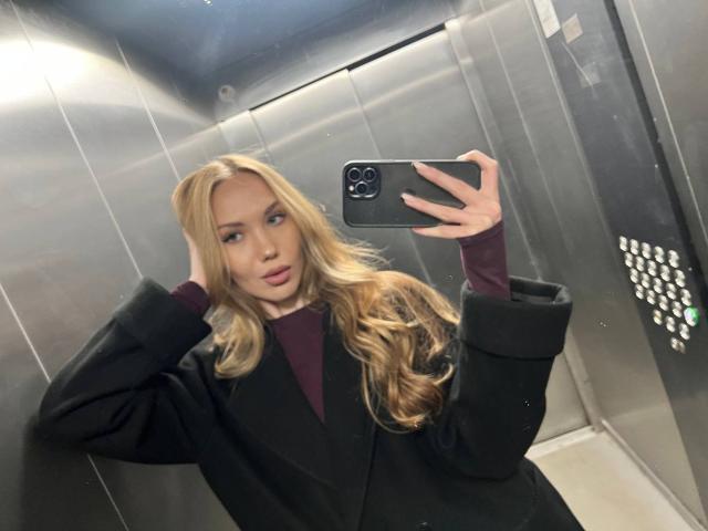 BrettRannels with long blonde hair, wearing a black blazer and maroon top, taking a mirror selfie in a metal elevator, holding a black iPhone. Boobs, ass, tits not visible.