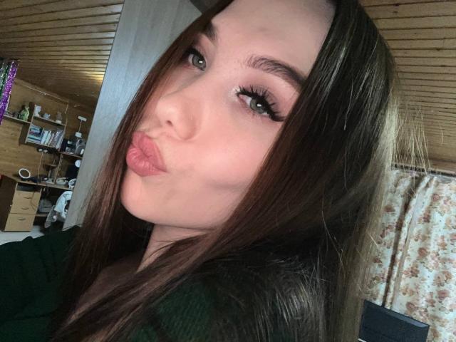 BrettRannels with pouty pink lips, long brown hair, green eyes, and thick eyelashes, wearing a green top, in a wooden room.