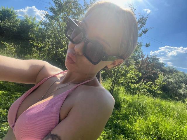 BrettRannels with short blonde hair, wearing black sunglasses, pink ribbed bikini top, pouting lips, and sunlight flaring in the background. Her tits are noticeable. Green grass and trees.