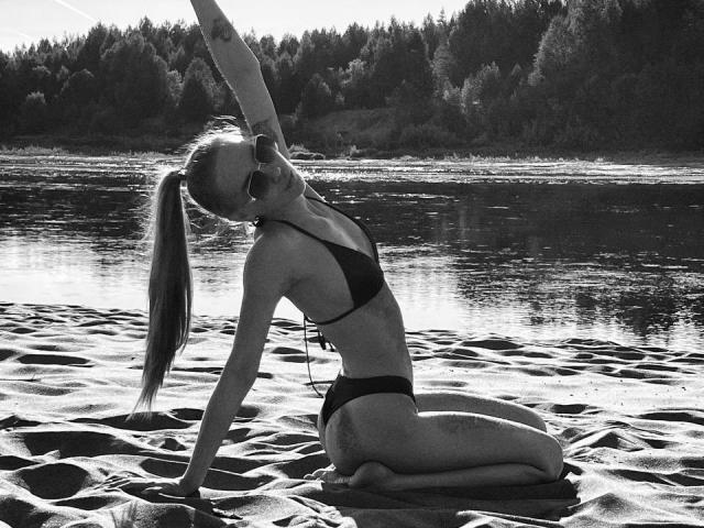 BrettRannels with long ponytail, wearing a black bikini, kneeling on sandy beach, stretching arm overhead, showing toned ass and small tits, in black-and-white photo.