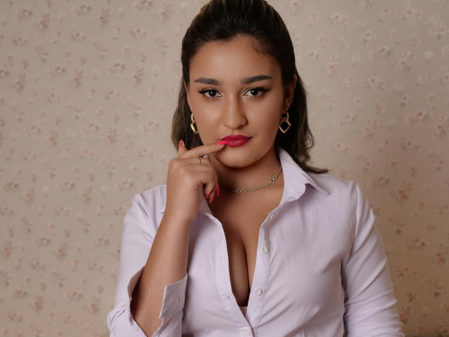 GoldenHear with brown hair, pink lipstick, and white shirt, touching her red-painted finger to her lips, showing her tits, wearing gold earrings, and a chain necklace.