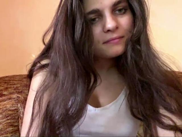 Jinny Kretschman, avec ses longs cheveux bruns ondulés, son débardeur blanc et son sourire discret, regarde légèrement sur le côté, montrant de petites seins et un visage mignon..