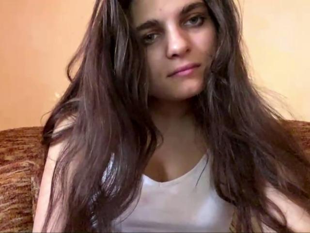 Jinny Kretschman, con lunghi capelli castani ondulati, indossa una canotta bianca che mette in mostra piccoli seni e ha un sorriso leggero e rilassato. È seduta su un divano marrone con una texture particolare..