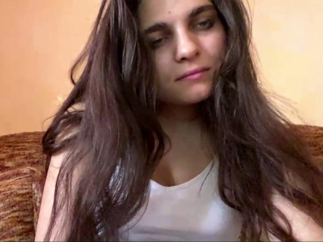 Jinny Kretschman, con cabello castaño largo, vestida con una camiseta blanca, mostrando sus pequeños senos, mirando hacia abajo con una ligera sonrisa, sentada en un sofá marrón con textura..