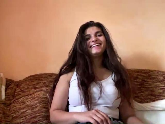 JinnyKretschman with long brown hair, white tank top, smiling, sitting on a brown patterned couch, hands on lap, cute, medium tits, light skin, casual, relaxed.