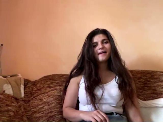 JinnyKretschman tem cabelos castanhos longos, uma camiseta branca e está sentada em um sofá marrom, com um sorriso discreto, parecendo relaxada. Seus seios são pequenos e bonitos..