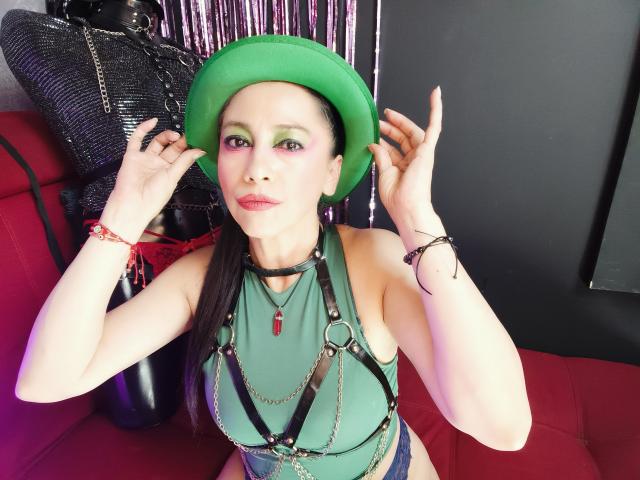 NoemiPrettiest with green hat, green halter top, black harness, and pink makeup, holding hat, showing off tits, on red couch, black wall, and shiny black bag in background.