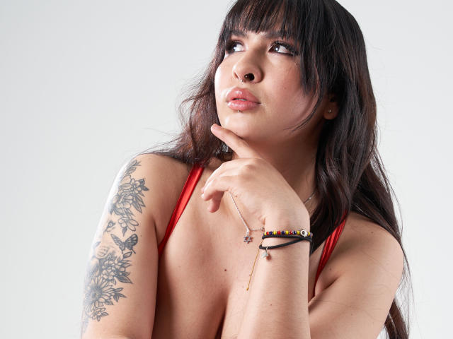 CelesteNeirafg with dark hair, red top, tattoos on right arm, thinking with finger on chin, wearing bracelet, looking up, pouty lips, pale skin, light makeup, soft light.
