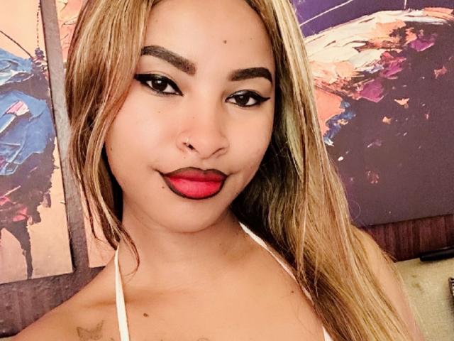 Plaissir69 has tan skin, long blonde hair, dark eyebrows, black eyeliner, red lipstick, and a small nose ring, wearing a white halter top, smiling slightly.
