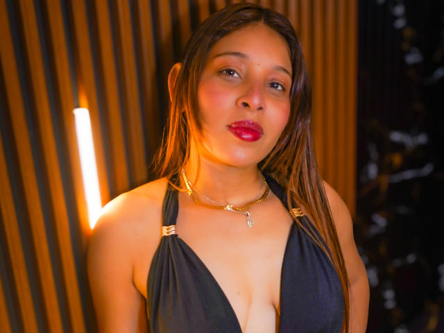 JaneKetty with wet brown hair, red lipstick, and a deep black halter top showing her big boobs and cleavage. She wears a gold chain with a small pendant.