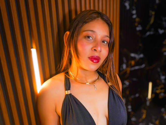 JaneKetty with brown hair, pink lipstick, and a deep black dress showing her tits, wearing a gold necklace, in a warm-lit, wooden background.