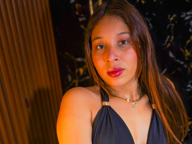JaneKetty with brown hair, red lips, black halter top, gold necklace, and gold earrings, has a cute face and small tits, looking directly at the camera.