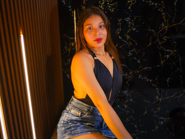 JaneKetty with long brown hair, red lipstick, black halter top, and blue denim skirt, sitting, showing her ass, against a dark marble wall, illuminated by warm light.