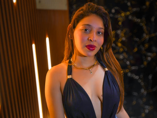 JaneKetty with tan skin, long brown hair, red lipstick, wearing a deep blue halter dress, showing her tits, gold necklace, standing in a warmly lit, wooden, and dark background.