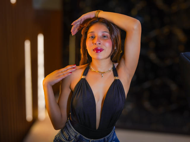 JaneKetty with brown hair, red lipstick, black halter top showing her tits, arm raised, hand on chest, gold necklace, blue jeans, confident smile, warm light, indoor setting.